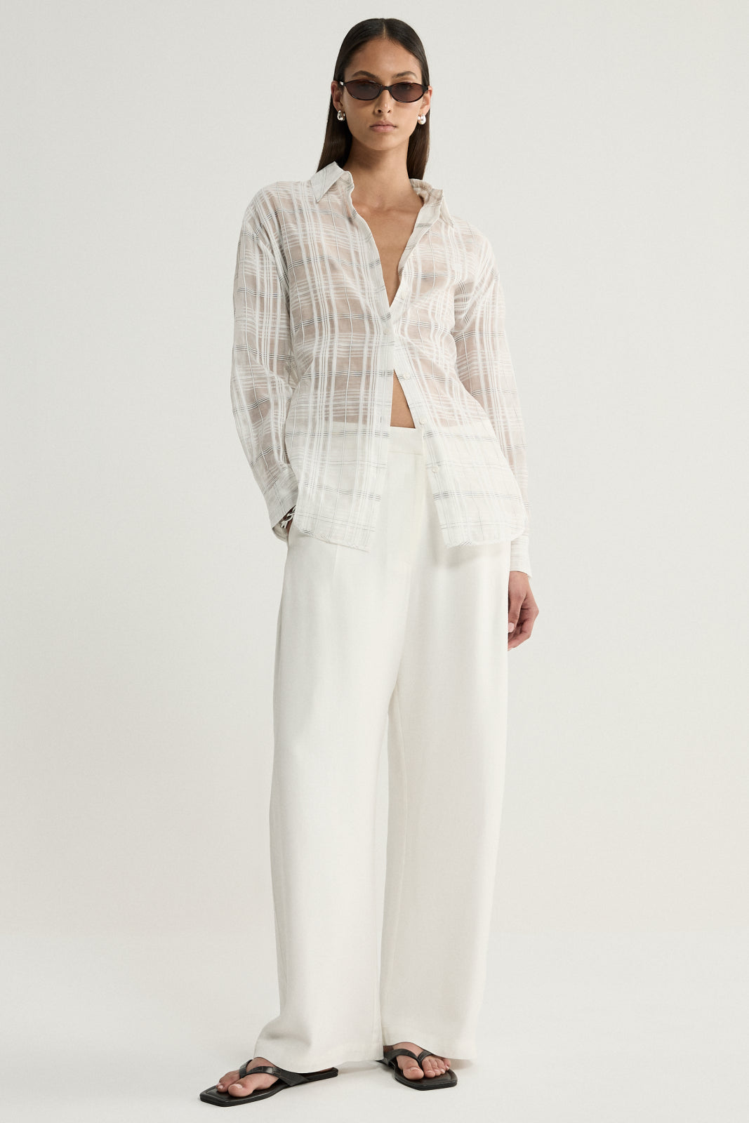 A model wearing a white semi-sheer checked button-up shirt with an open collar and relaxed sleeves, styled with ivory wide-leg trousers and black sandals in a straight-on studio pose.