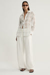 A three-quarter view of a model wearing a white check shirt with a collared neckline, button front and softly gathered sleeves, paired with ivory trousers and dark sunglasses.