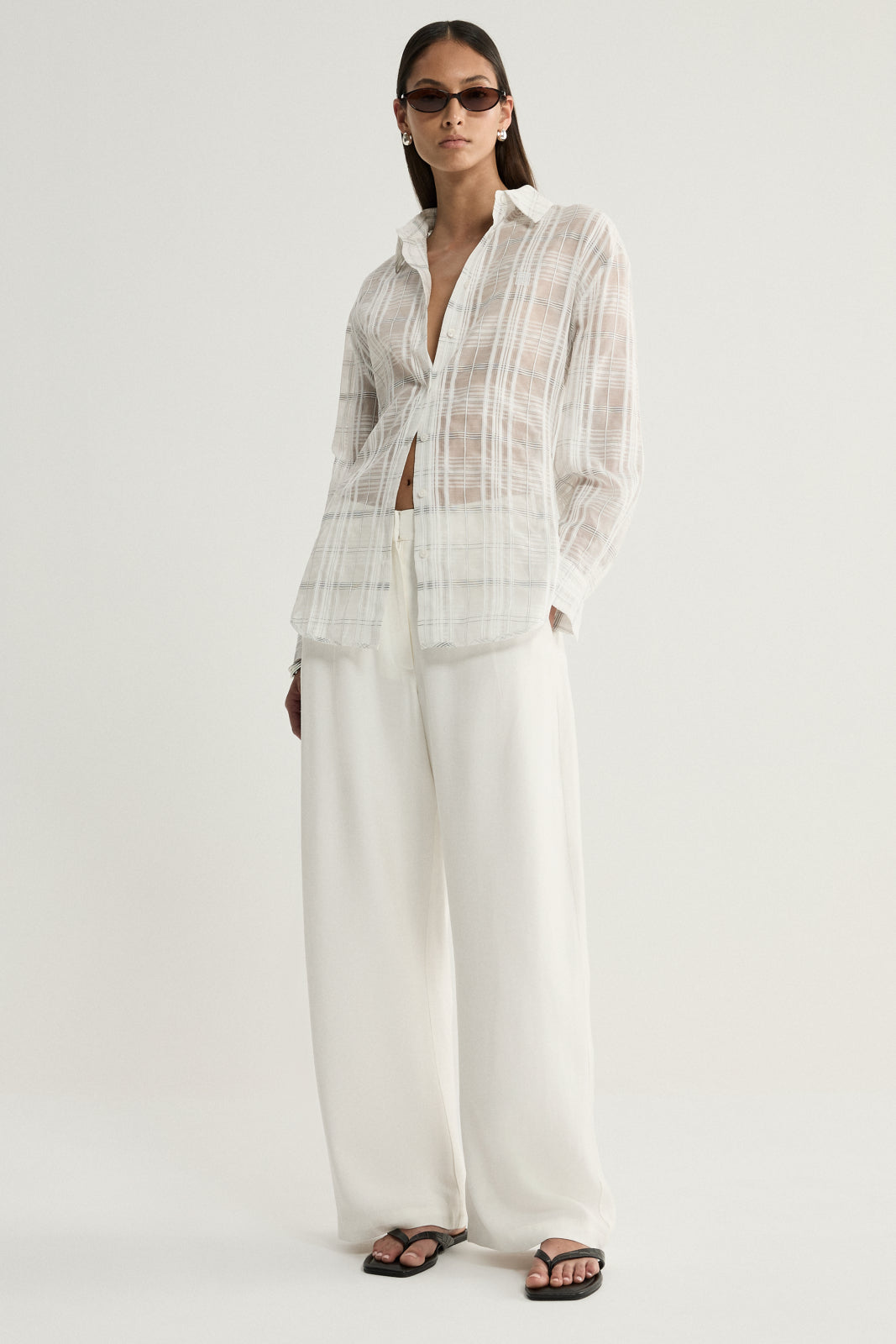 A three-quarter view of a model wearing a white check shirt with a collared neckline, button front and softly gathered sleeves, paired with ivory trousers and dark sunglasses.