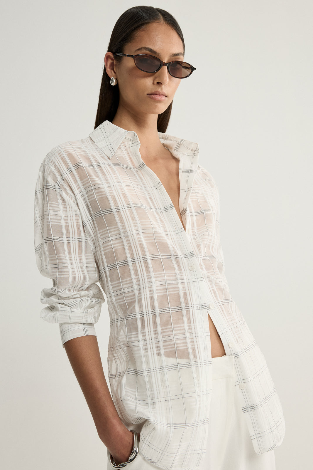 A model wearing a white semi-sheer check shirt with a relaxed cut, open front neckline and long sleeves pushed up to the forearms, showing the shirt's length and neutral checked pattern.
