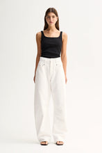 A woman modeling a black square-neck tank top and high-waisted, wide-leg white trousers against a plain white studio background.