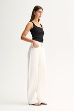 A woman modeling a black sleeveless tank top and high-waisted, wide-leg white trousers against a plain white studio background.