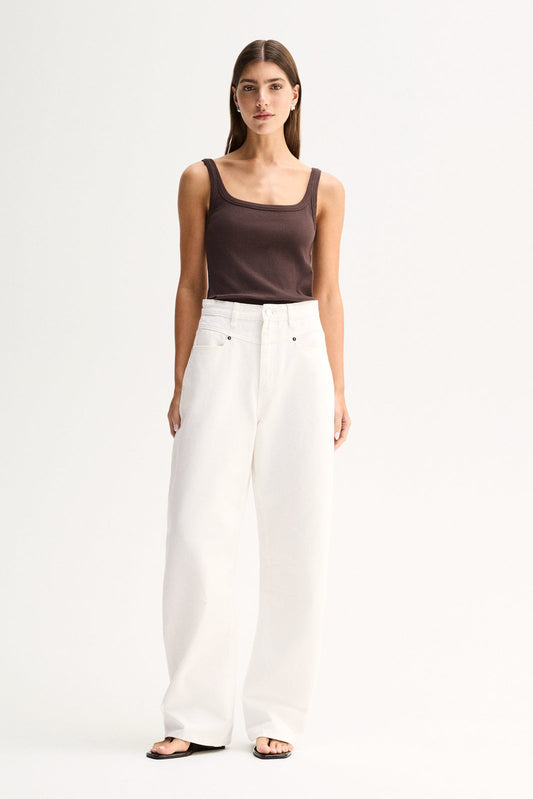A woman wearing a chocolate fitted ribbed tank with a low scoop neckline and thin shoulder straps, paired with wide-leg white pants.