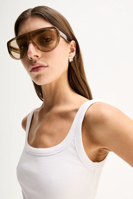 A close-up of a woman wearing oversized square sunglasses with thick brown frames and brown-tinted lenses, paired with a white ribbed tank top against a plain light background.