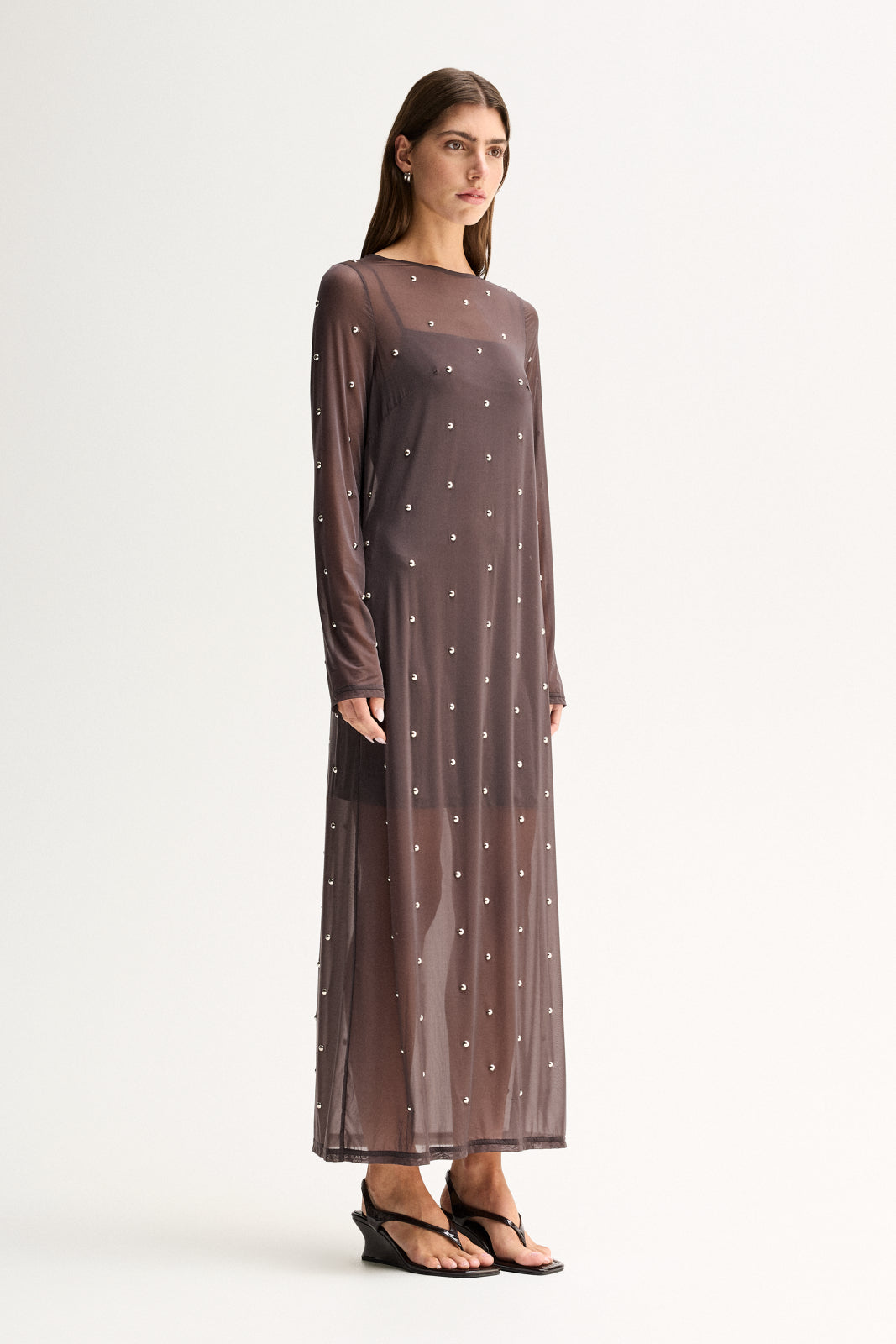 Side view of a woman wearing a slate sheer long-sleeve maxi dress with scattered stud details and a flowing full-length skirt.