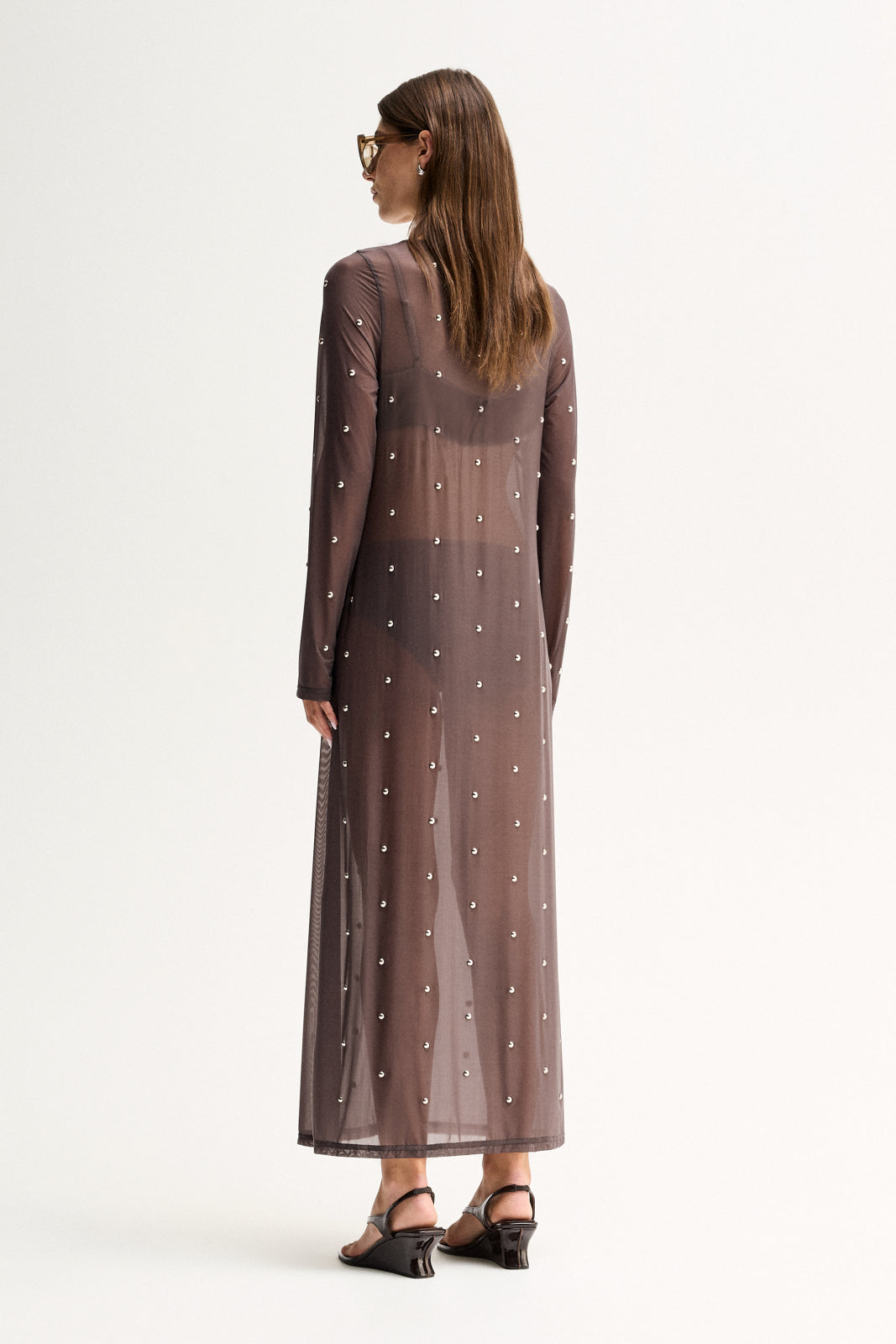 Back three-quarter view of a woman wearing a slate sheer maxi dress with long sleeves and scattered stud detailing.