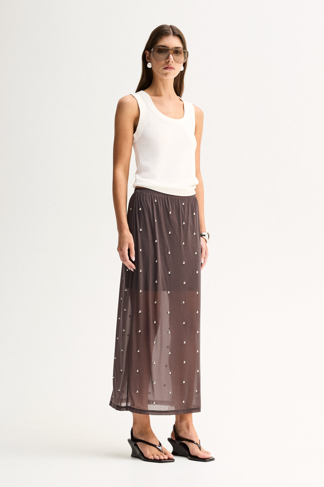 Three-quarter view of a woman wearing a slate sheer midi skirt with scattered stud details and a white sleeveless top.