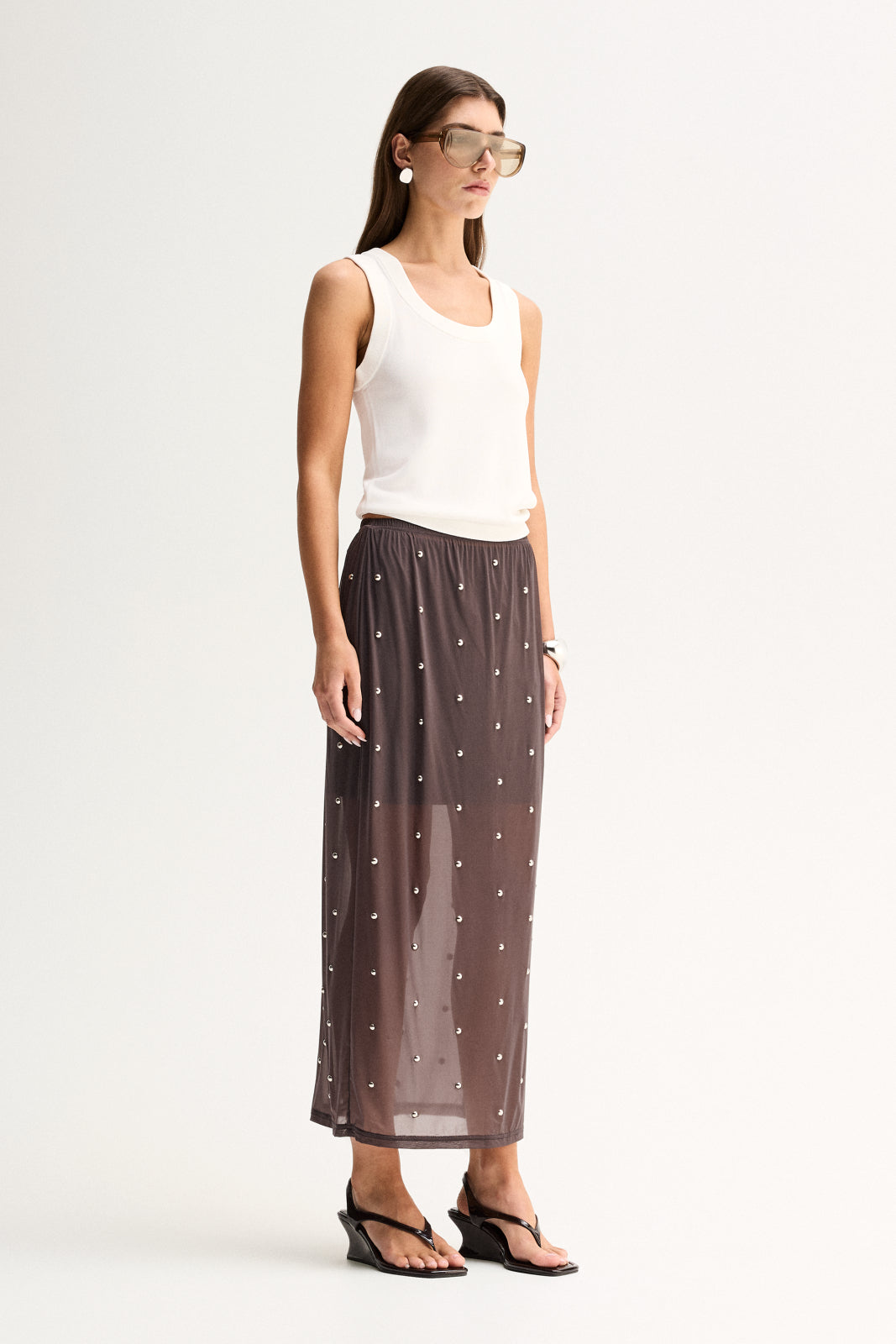 Side view of a woman wearing a slate sheer midi skirt with an elasticated waist, stud detailing and a flowing midi-length hem.