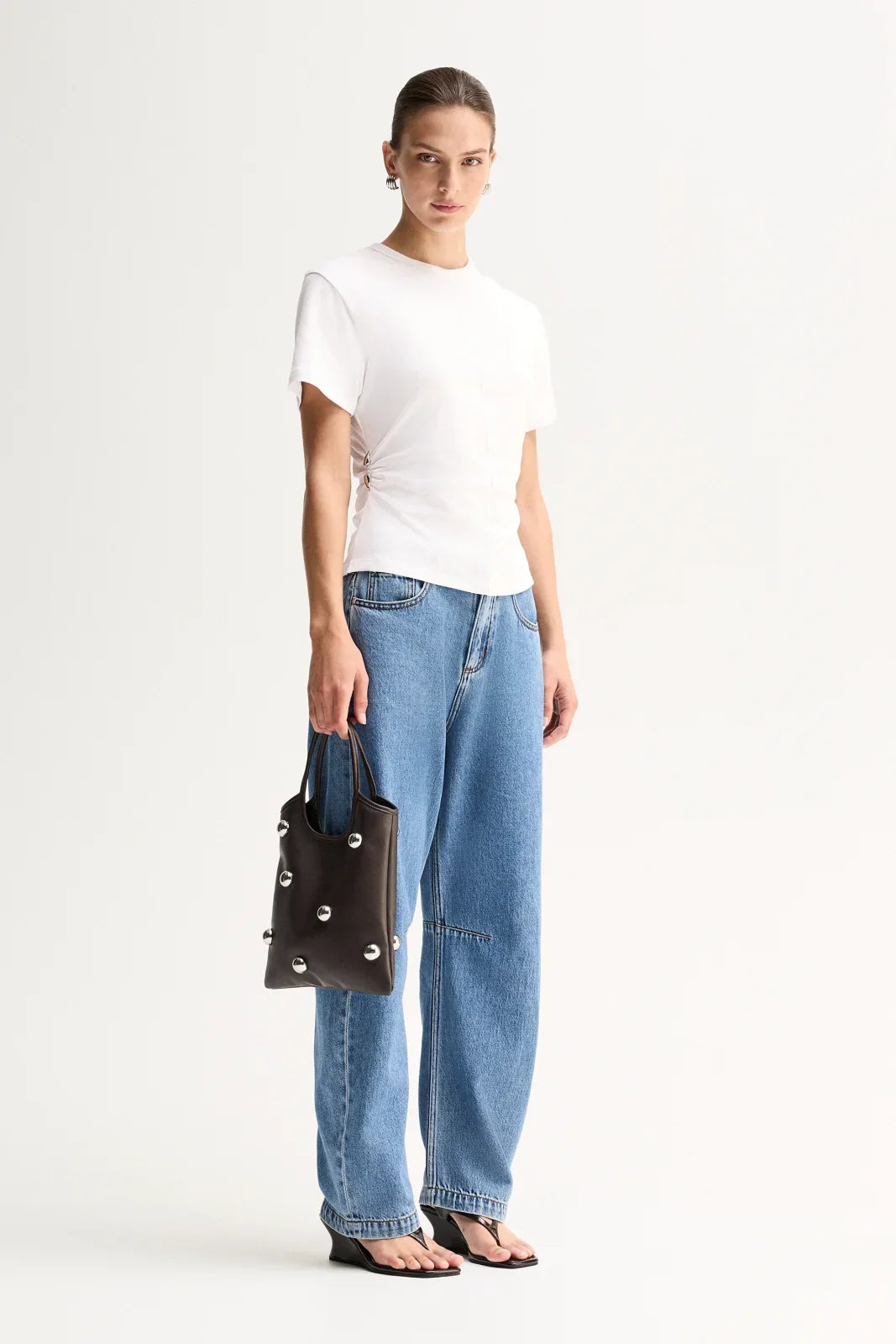 A model wears the white side-gathered top with blue wide-leg jeans while holding a black studded bag in an angled full-length view.