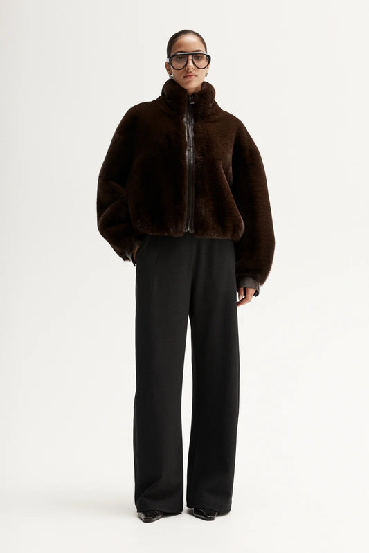 Model wearing a dark brown plush cropped jacket with black wide-leg trousers and oversized glasses, showing the high collar and relaxed silhouette, front view