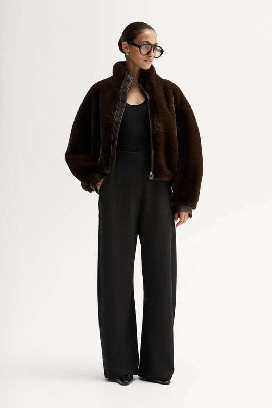 Model wearing the dark brown plush cropped jacket open over a black top with black wide-leg trousers and oversized glasses, showing the soft textured finish, front view