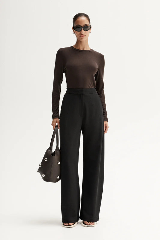 Woman wearing a brown long-sleeve top and black high-waisted pants, holding a black handbag on a white background.
