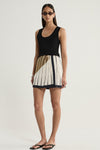 A model wearing a cream mini skirt with fine dark vertical stripes, a diagonal moss-coloured panel at the waist and a contrasting dark hem, styled with a fitted black tank top, narrow sunglasses and black flip-flops in a full-length front view.