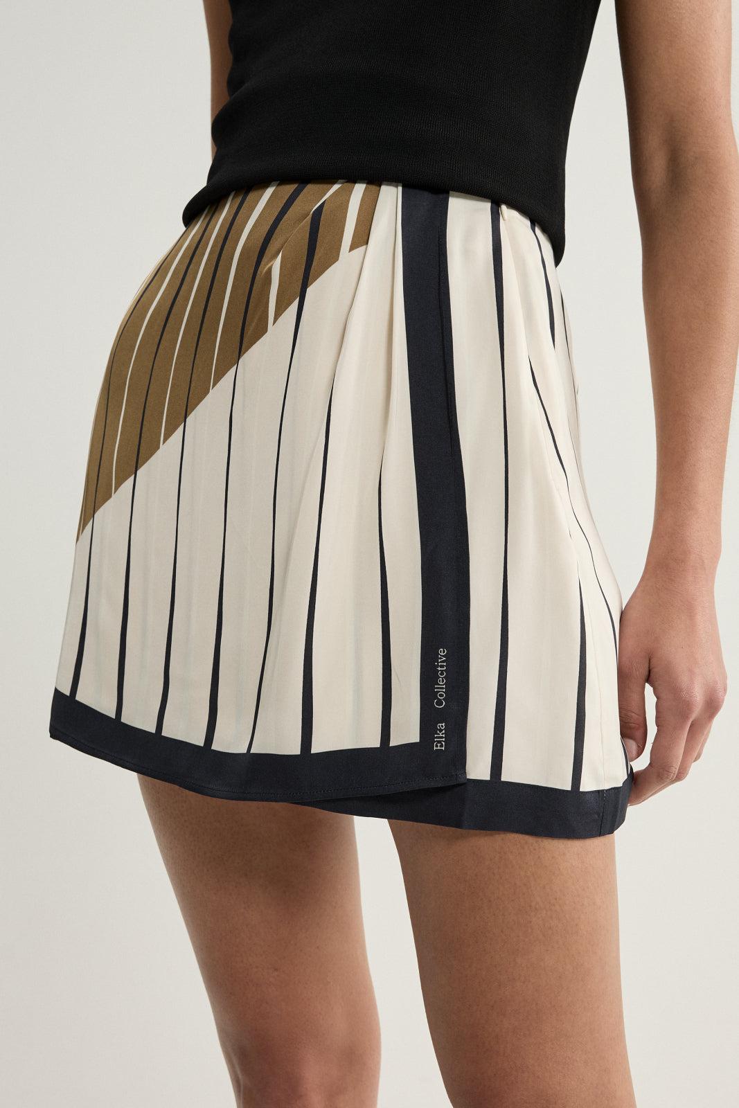 A close-up of the cream mini skirt showing fine dark vertical stripes, a diagonal moss and brown-toned panel across the upper section, gathered fabric at the waist and a wide dark border along the hem.