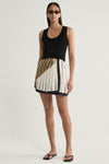 A model stands facing forward in a cream mini skirt with dark pinstripes, an asymmetrical moss-coloured panel and a dark hem, paired with a fitted black sleeveless top and black flip-flops against a plain studio background.