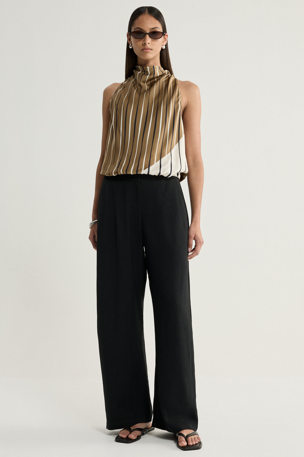 A model stands facing forward in a moss, cream and dark striped sleeveless top with a high neckline, tucked into wide-leg black trousers and paired with black sandals, showing the overall silhouette and full-length styling against a plain studio background.