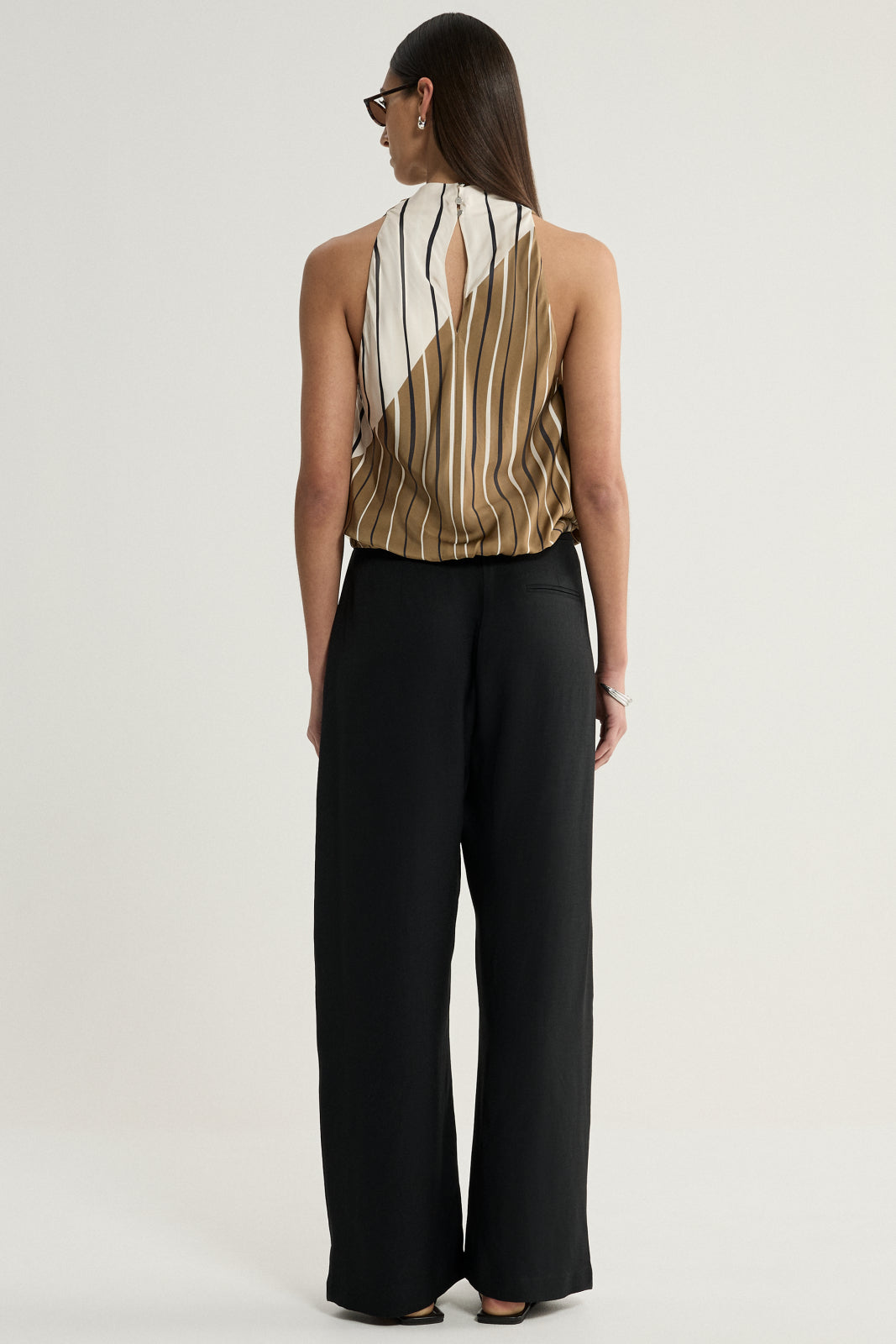 A rear view of a model wearing the striped sleeveless top with black wide-leg trousers, showing the high neckline, open upper-back detail with a narrow fastening, and the contrasting cream and moss sections of the print.