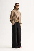 Model wearing the Camel Elka Collective Trixie Jacket zipped up with black wide-leg trousers, showing the funnel neck and cropped silhouette, full-length angled view