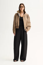 Model wearing the Camel Elka Collective Trixie Jacket open over a black top with black wide-leg trousers, showing the funnel neck and elasticated hem, full-length front view