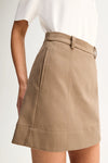A close-up of a model wearing a beige A-line mini skirt with a matching belt and a white top, against a plain light background.