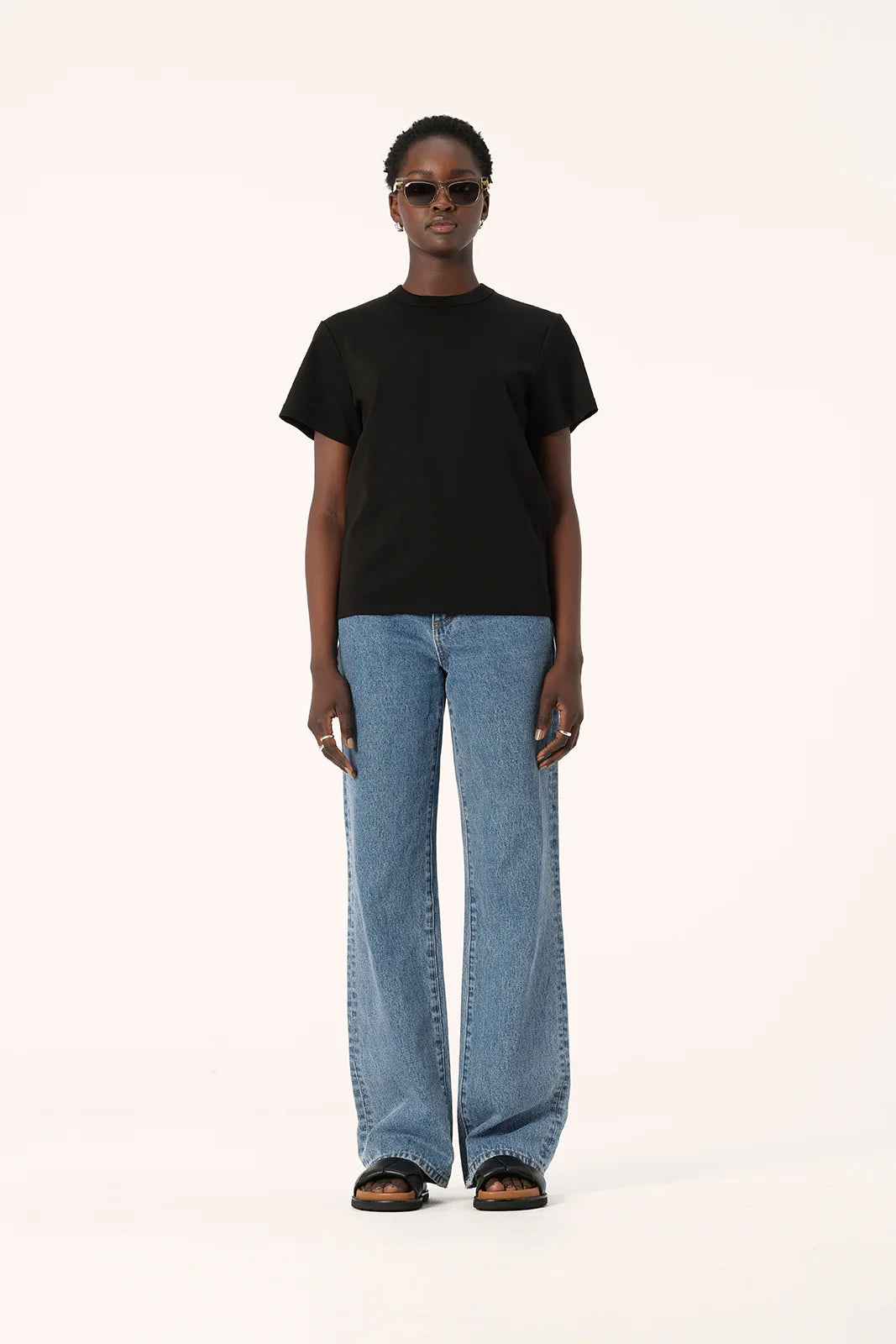 Model wearing the black Zoe Tee with light blue straight-leg jeans, black sandals and sunglasses, showing the crew neckline, short sleeves and slightly oversized boxy fit, full-length front view
