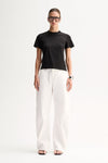 A model wearing a black short-sleeve T-shirt with a round neckline and relaxed silhouette, paired with white wide-leg trousers and black flat sandals.