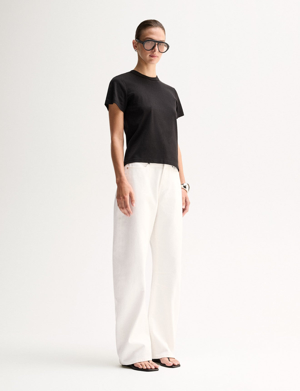 An angled front view of a model wearing a black crew-neck T-shirt with short sleeves and a relaxed shape, styled with white trousers and black sandals.