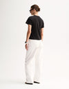 A rear three-quarter view of a model wearing a black T-shirt with short sleeves and a plain back, paired with white trousers and black flat sandals.