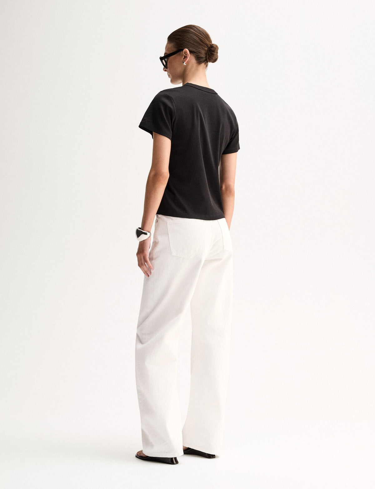 A rear three-quarter view of a model wearing a black T-shirt with short sleeves and a plain back, paired with white trousers and black flat sandals.
