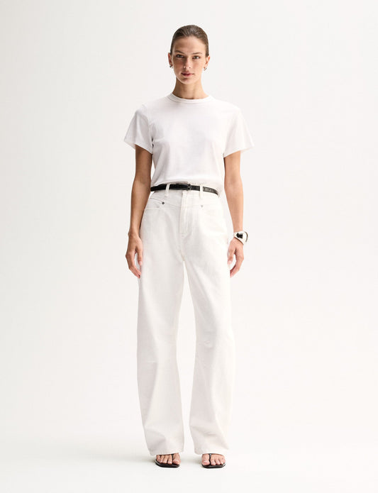 A model wearing a white relaxed-fit T-shirt with a round neckline and short sleeves, tucked into white straight-leg trousers with a black belt and black flat sandals.