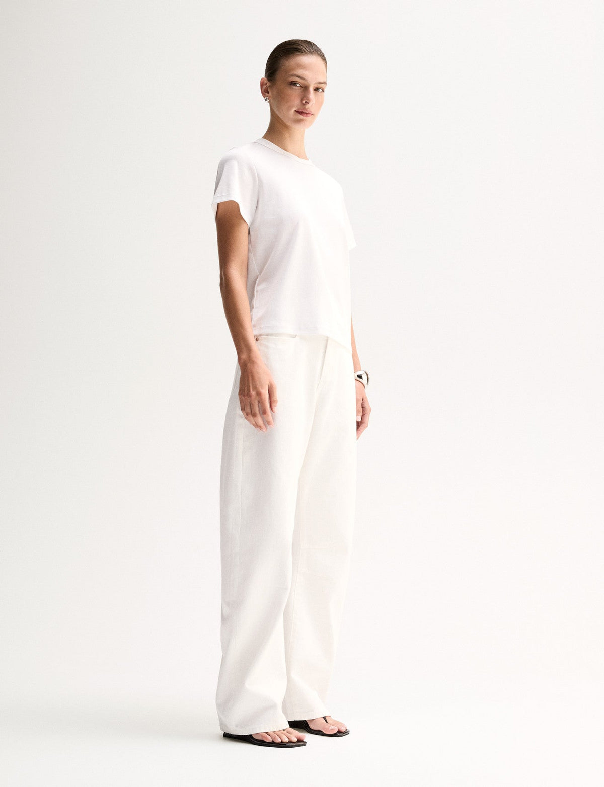A side view of a model wearing a white short-sleeve T-shirt with a classic crew neckline and relaxed shape, paired with white trousers and black flat sandals.