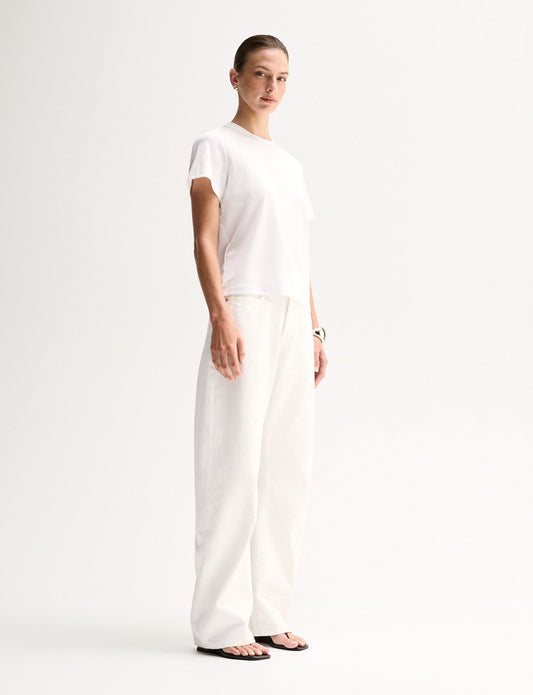 A side view of a model wearing a white short-sleeve T-shirt with a classic crew neckline and relaxed shape, paired with white trousers and black flat sandals.