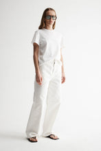 Model wearing the white Zoe Tee with white trousers and flat sandals, showing the relaxed silhouette, short sleeves and straight hem, full-length front angled view
