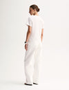 A rear three-quarter view of a model wearing a white relaxed T-shirt with short sleeves and a simple back, styled with white straight-leg trousers and black sandals.