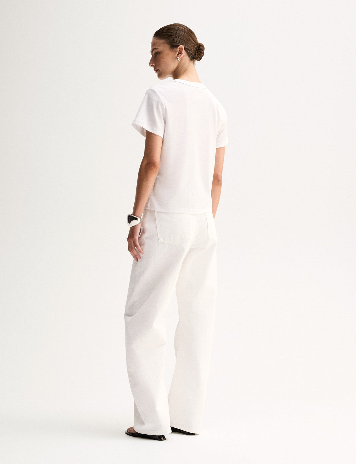 A rear three-quarter view of a model wearing a white relaxed T-shirt with short sleeves and a simple back, styled with white straight-leg trousers and black sandals.