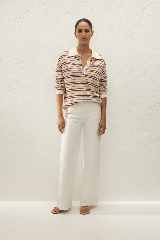 Full-length front view of the striped oversized polo styled with white wide-leg trousers and neutral sandals against a textured cream wall.