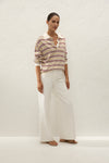 Three-quarter view of the relaxed striped polo styled with white trousers and sandals against a textured neutral background.