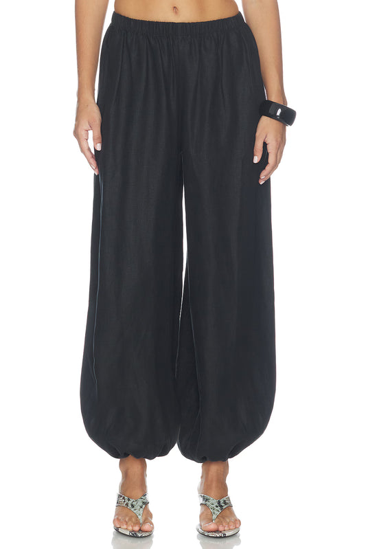 Close-up of black balloon pants showing the gathered elastic waistband, relaxed fit through the hips and legs, voluminous silhouette and gathered ankle cuffs, paired with snakeskin-patterned heeled sandals.