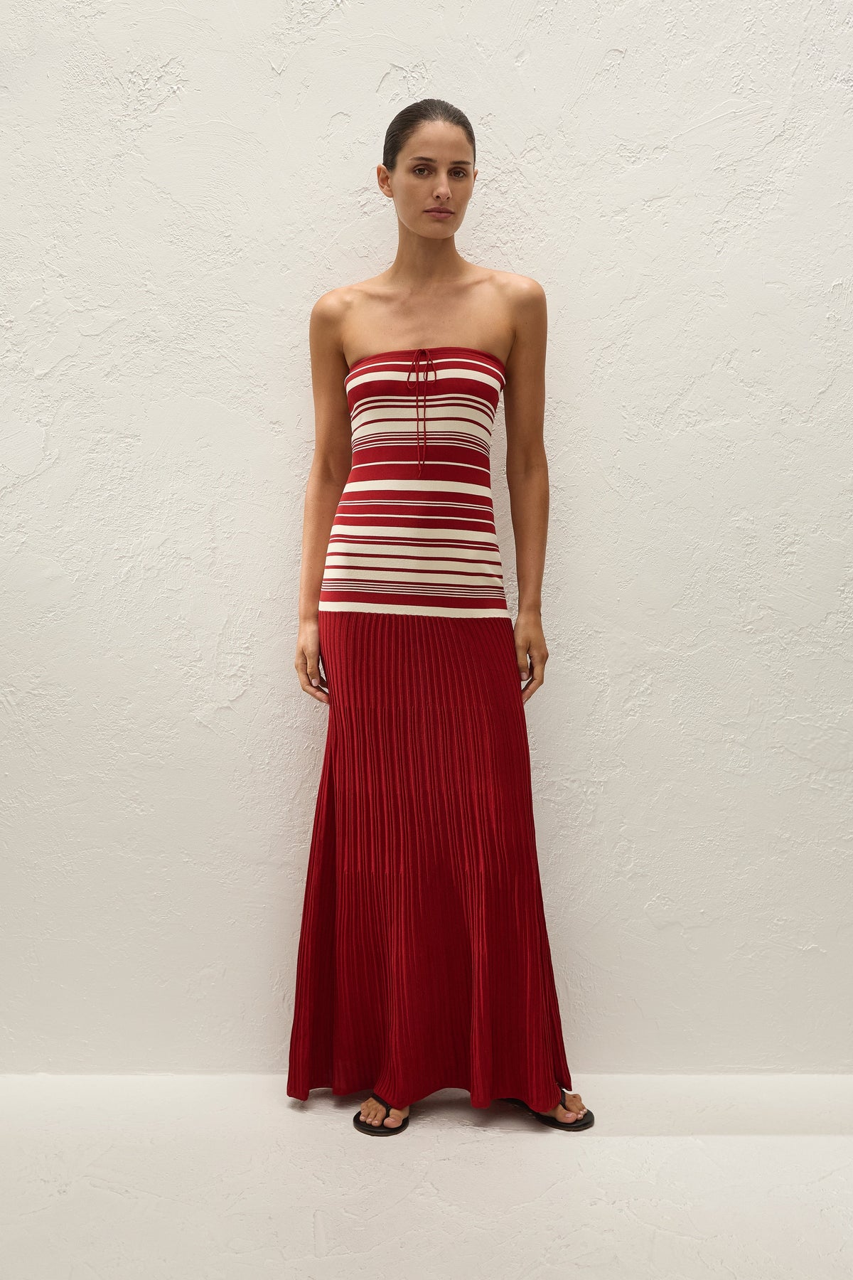A woman wears a red and cream striped strapless maxi dress with a long pleated red skirt.

