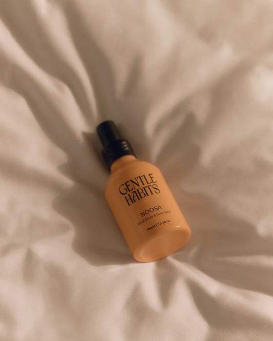 Gentle Habits Ritual Room & Linen Spray in Noosa shown lying on a light fabric surface with its rounded bottle and black spray top.