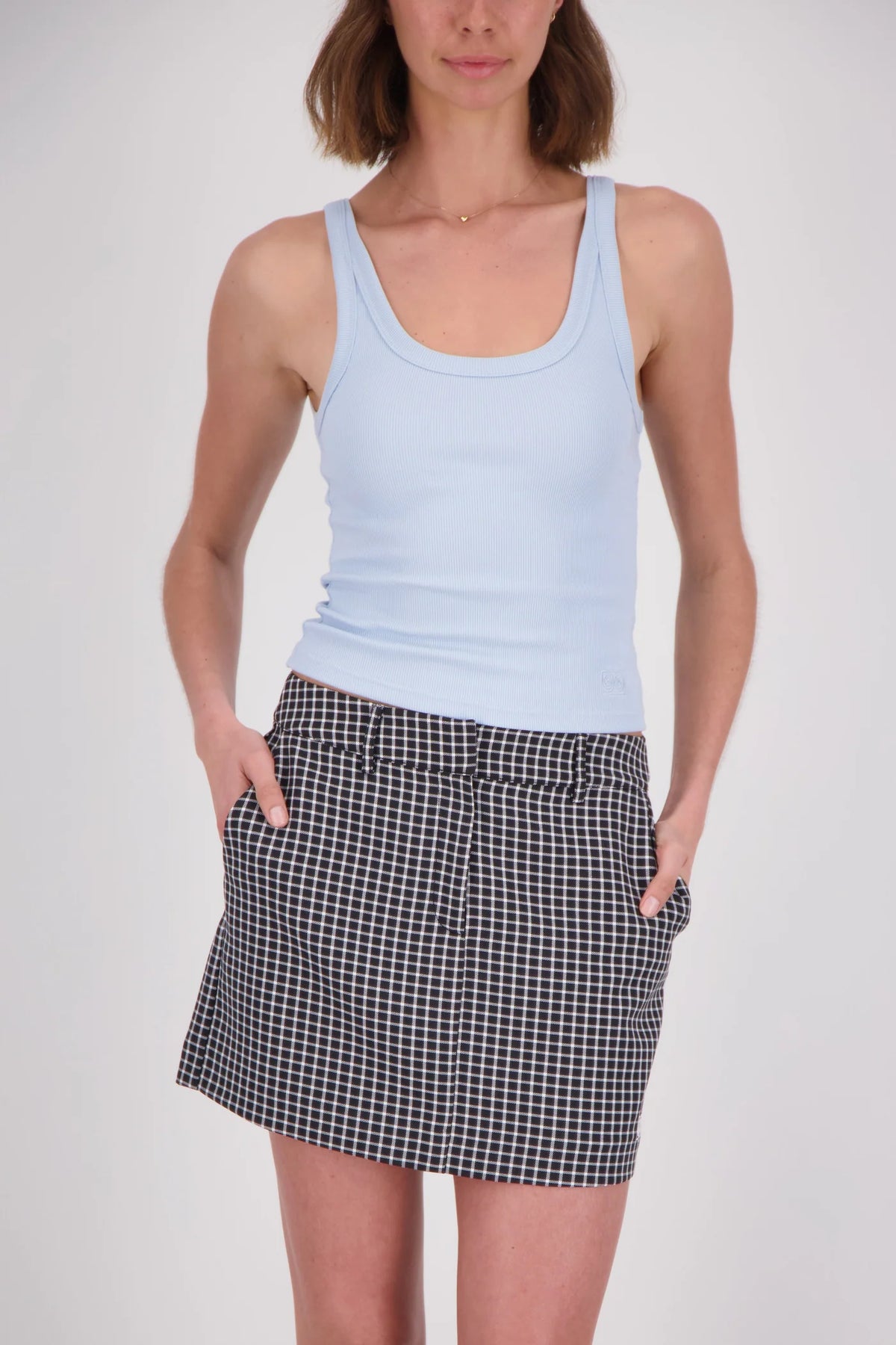 A woman wearing a pale blue ribbed tank top with a scoop neckline and wide shoulder straps, paired with a black-and-white checked mini skirt, photographed from the front with her hands in the skirt pockets.
