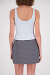 A rear view of a woman wearing a pale blue ribbed tank top, showing its wide shoulder straps, low scoop back and fitted shape, paired with a black-and-white checked mini skirt.