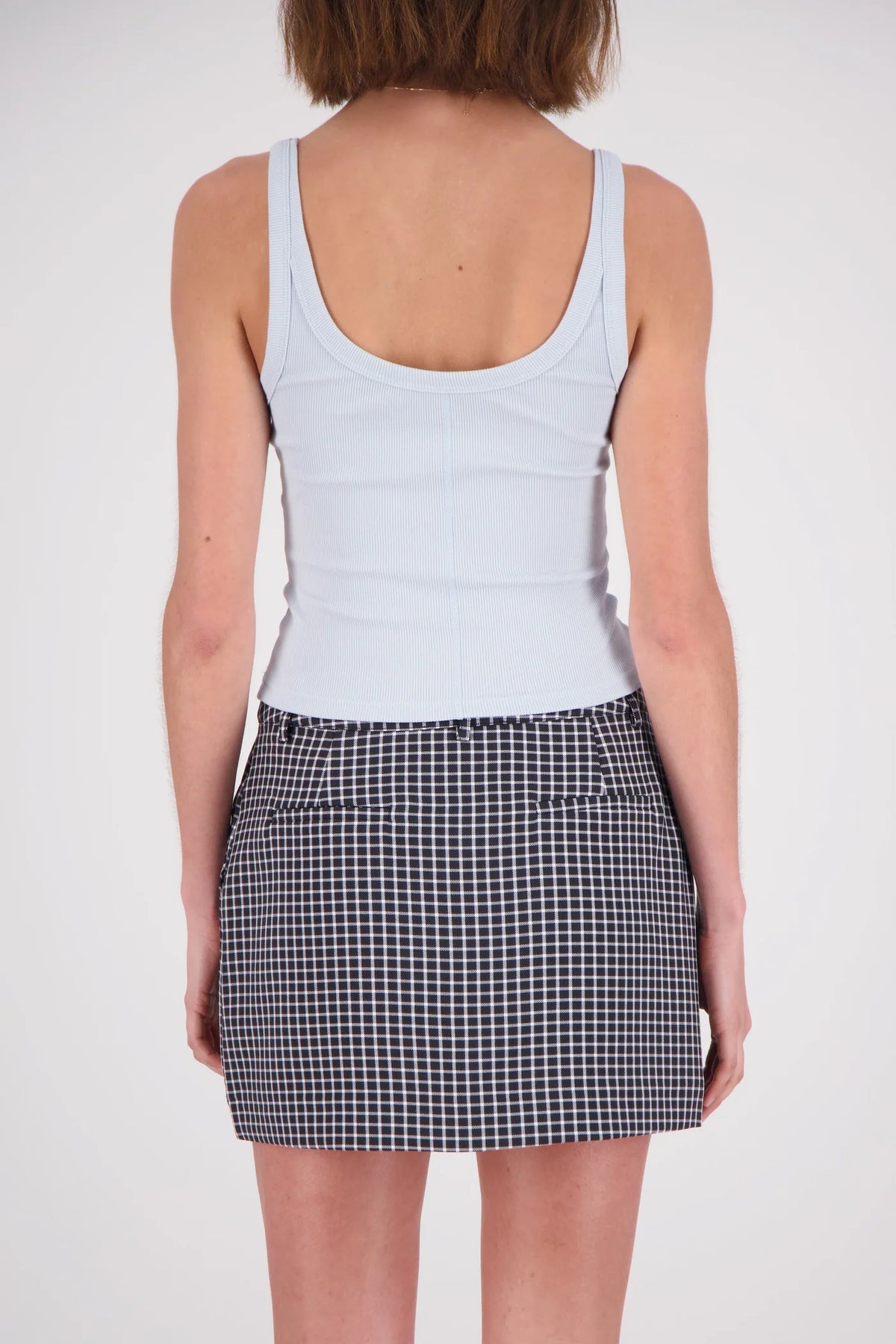 A rear view of a woman wearing a pale blue ribbed tank top, showing its wide shoulder straps, low scoop back and fitted shape, paired with a black-and-white checked mini skirt.