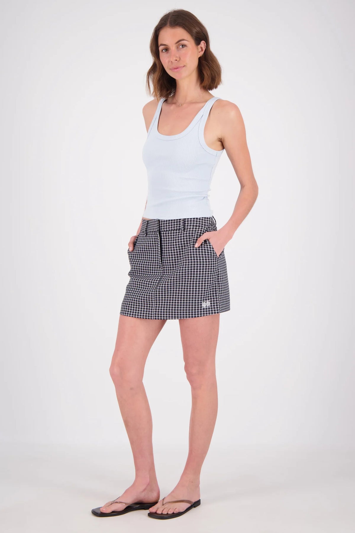 A full-length front view of a woman wearing a pale blue fitted ribbed tank top with a scoop neckline and wide shoulder straps, paired with a black-and-white checked mini skirt and flat sandals.