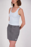 A woman wearing a pale blue ribbed tank top with a scoop neckline and wide shoulder straps, paired with a black-and-white checked mini skirt, shown from a three-quarter angle against a plain light studio background.