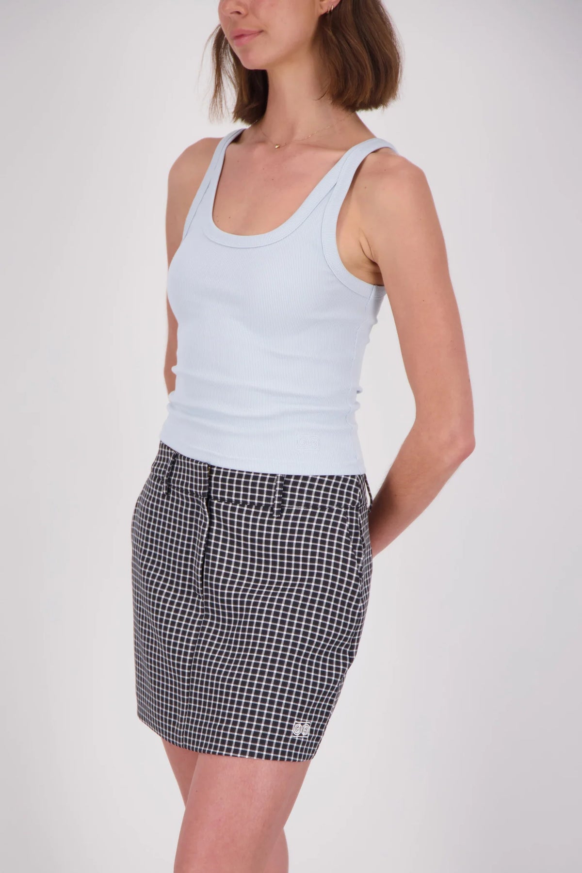 A woman wearing a pale blue ribbed tank top with a scoop neckline and wide shoulder straps, paired with a black-and-white checked mini skirt, shown from a three-quarter angle against a plain light studio background.