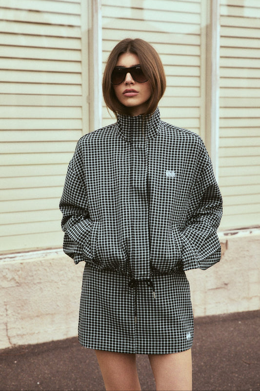  A model poses outdoors in a black and white check bomber jacket and matching mini skort, showing the jacket's relaxed silhouette, high neckline and coordinated check pattern.