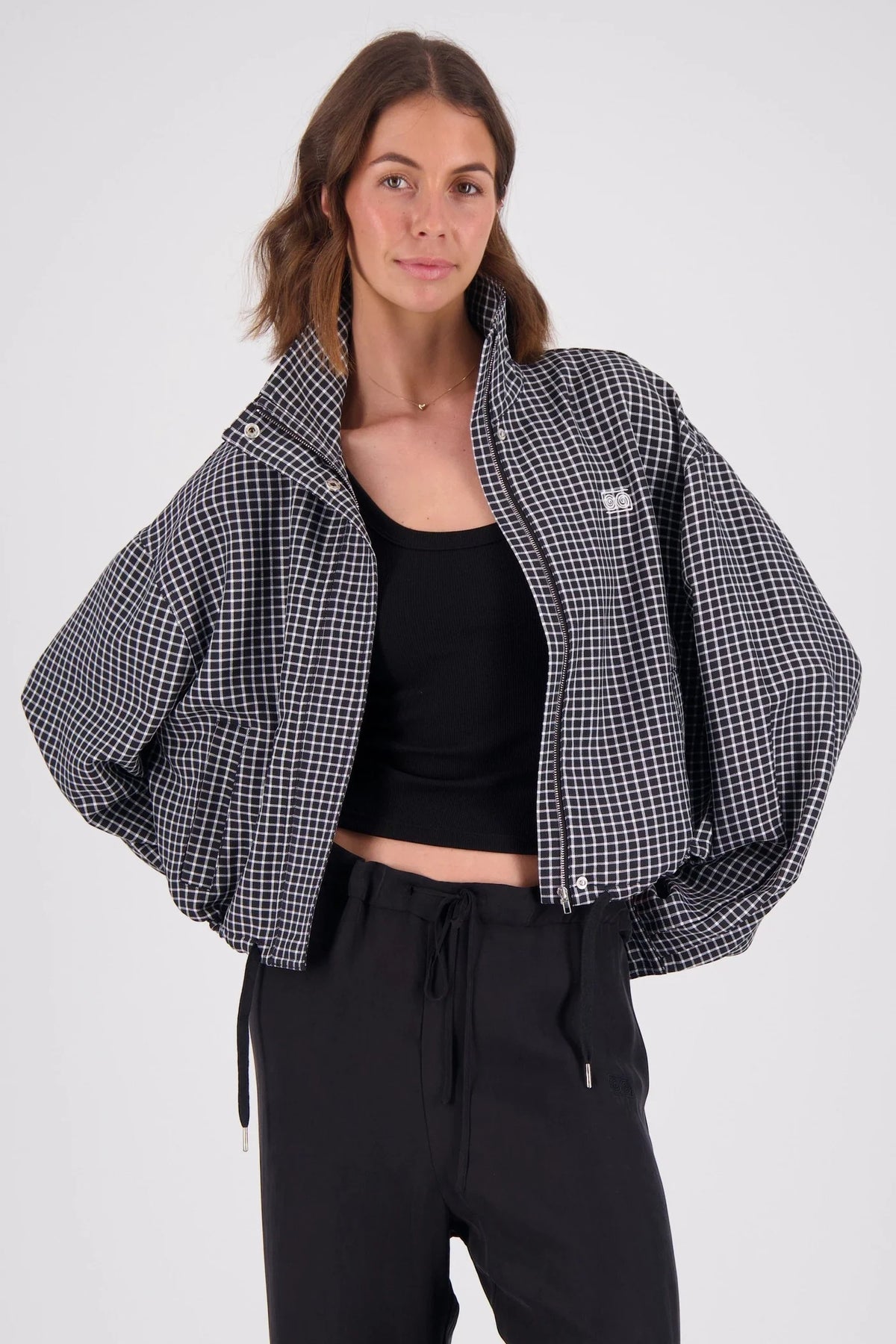 A model wears an open black and white check bomber jacket over a black cropped top and black drawstring trousers, showing the front zip, high collar, voluminous sleeves and relaxed cropped shape.