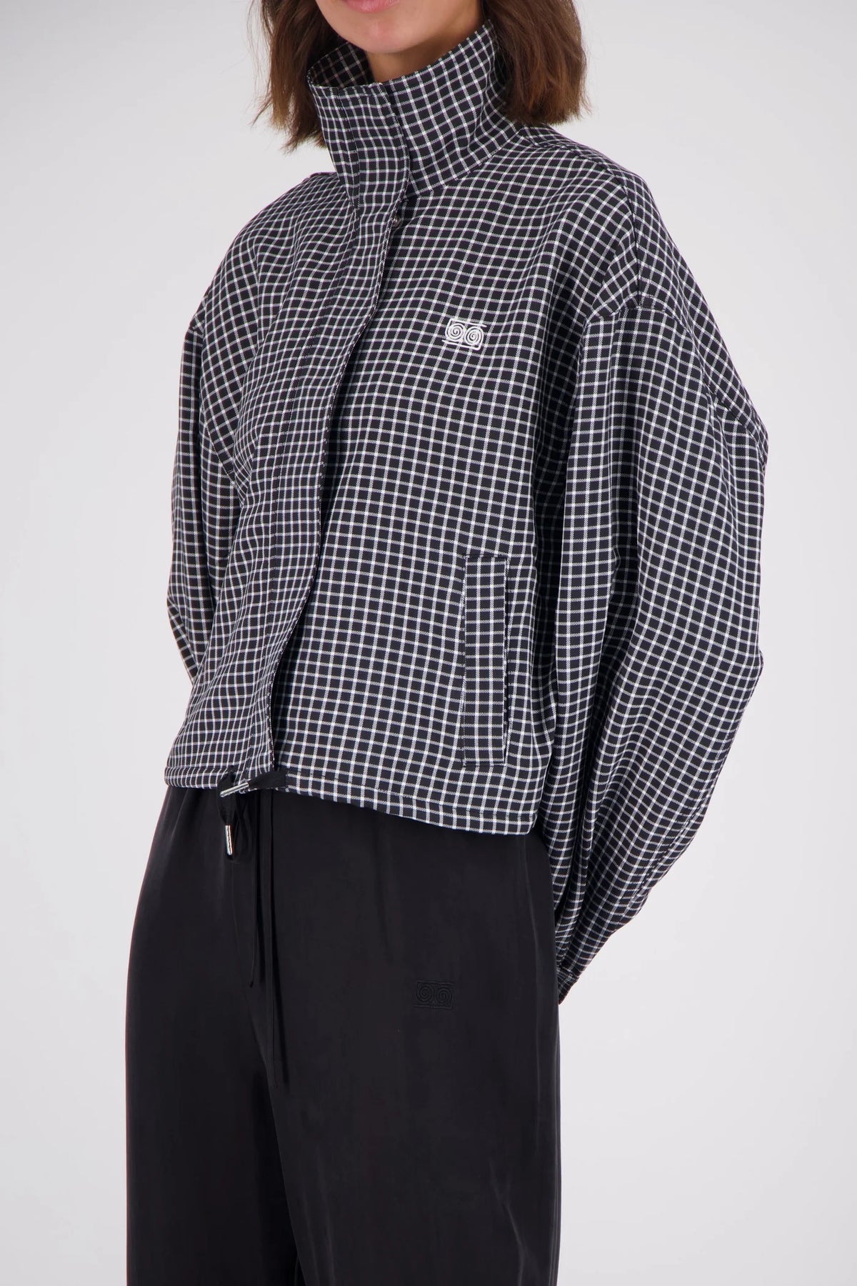 Close-up of a black and white check bomber jacket showing the raised collar, front zip fastening, chest logo embroidery and vertical front pocket.