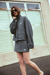 A model poses outdoors in a black and white check bomber jacket and matching mini skort, with one hand raised near the collar to highlight the long sleeves, relaxed fit and coordinated set.